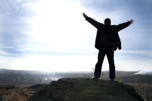 A person standing on a rock with their arms in the air, looking out over a vast landscape with a bright sky.