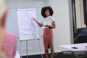 A woman with curly hair stands in front of a flipchart giving a presentation about how to make money on Facebook.