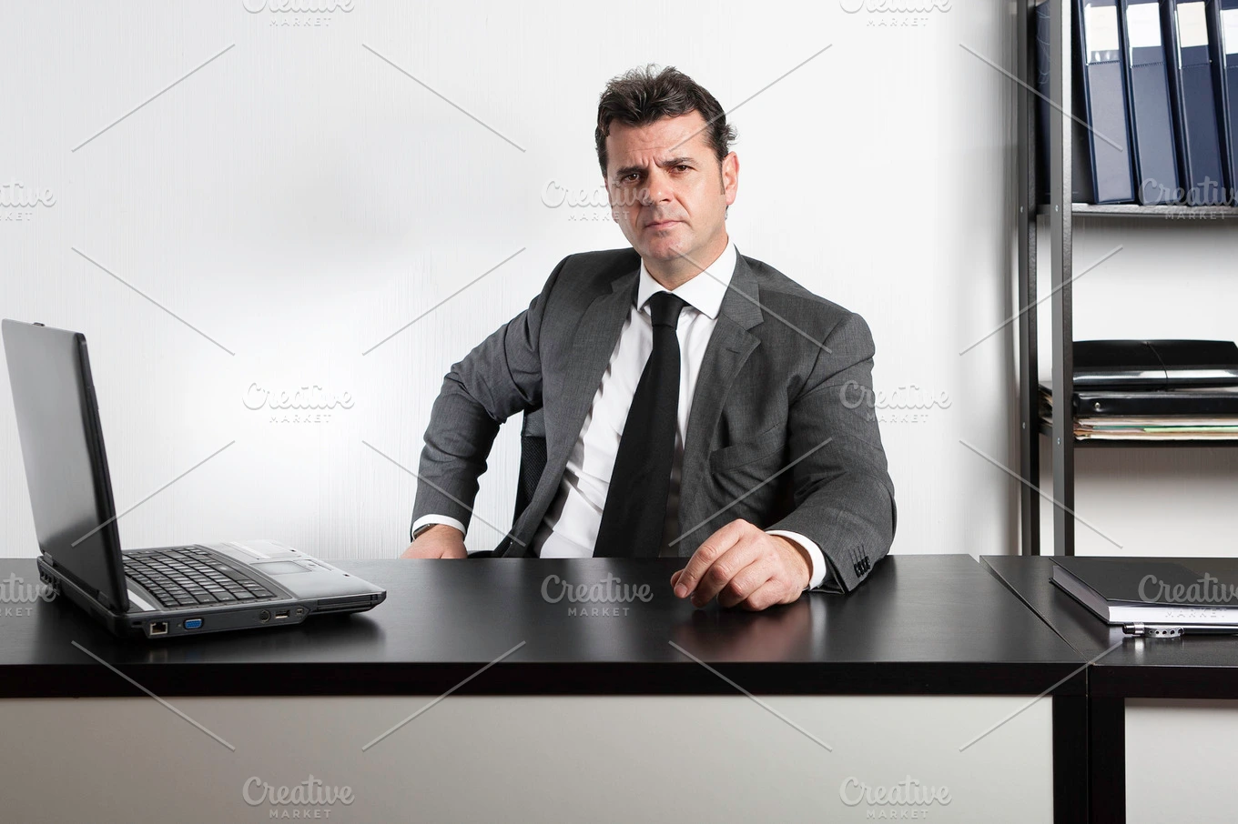 A businessman in a suit sits at his desk, looking focused and productive, with a laptop on the left side of the desk and a notebook on the right side.