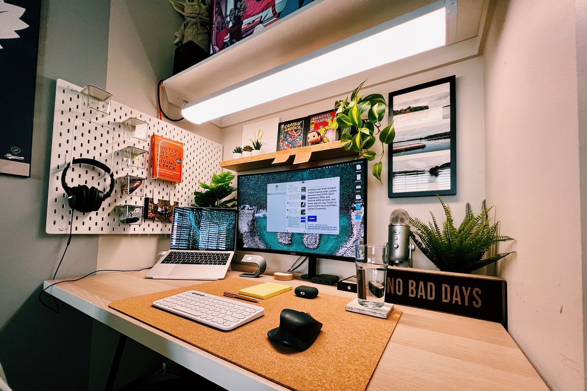 An organized office desk setup with natural light, an efficient L-shaped layout, minimal distractions, a corkboard with a pegboard, a desk lamp, plants, a framed print, a glass of water, a coaster, a keyboard, a mouse, a laptop, a monitor, headphones, a microphone, and a speaker.