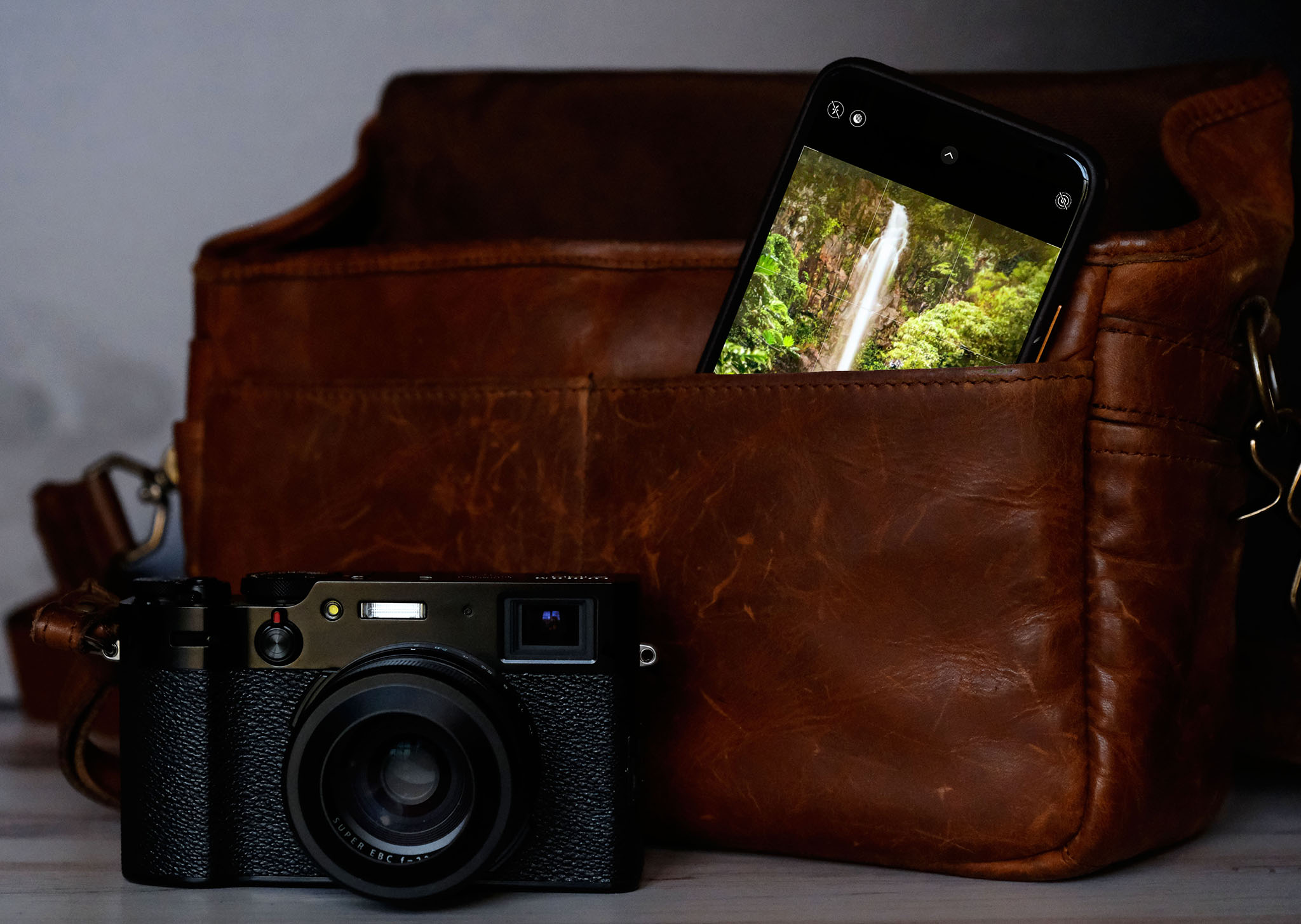 A photo of a camera and a smartphone in a brown leather bag with a photo of a waterfall on the screen.