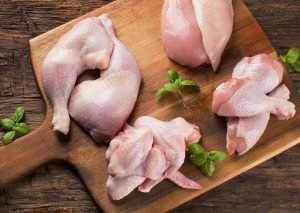 On a wooden cutting board are raw chicken parts including two drumsticks, two thighs, a half of a chicken breast, and two chicken wings with green basil leaves.