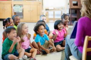 A group of children are sitting on the floor in a classroom. They are all smiling and looking at the teacher, who is sitting in front of them and reading a book.