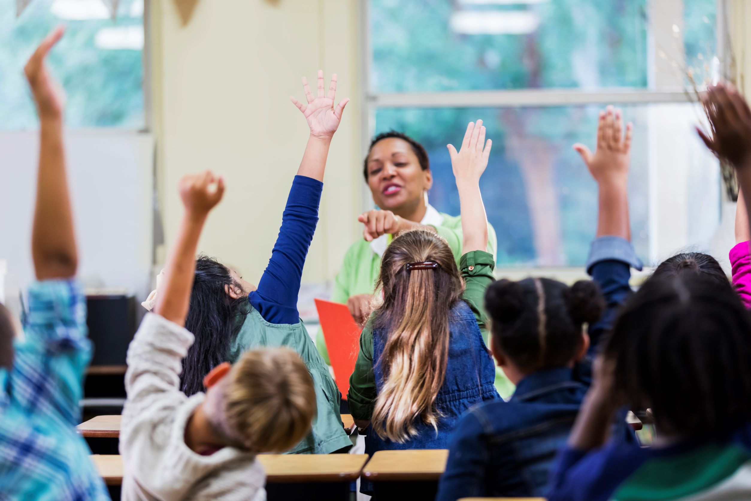 A classroom scene with children sitting at their desks raising their hands while the teacher stands at the front of the room smiling.