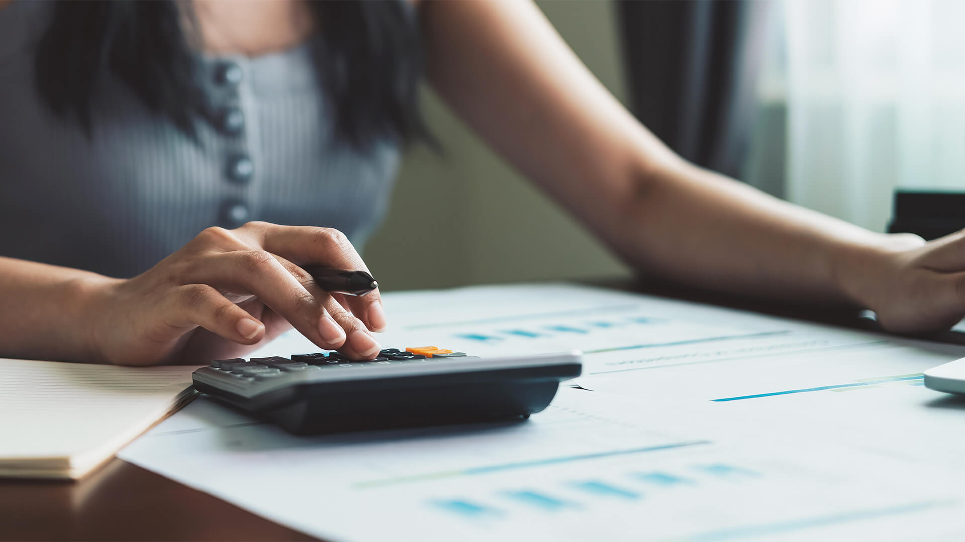 A person is using a calculator to manage their finances while looking at a computer screen and some papers with graphs on them.