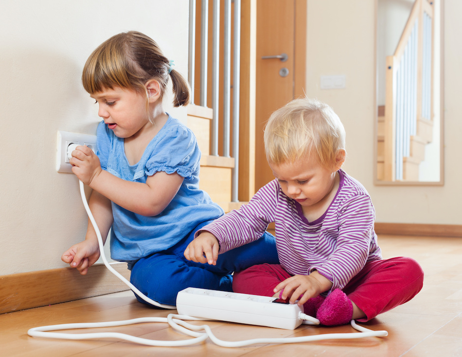 Two young children play with an electrical outlet and a power strip, illustrating the need to prevent home accidents.
