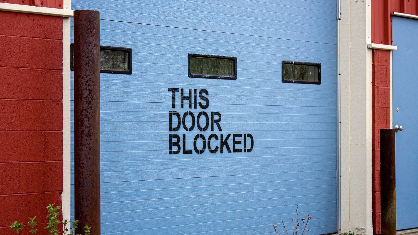 A blue garage door with the words 'This door blocked' spray-painted on it, in capital letters.