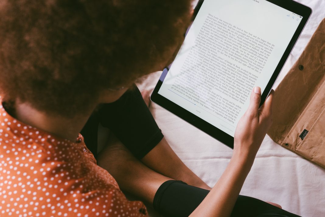 A person wearing a brown and white polka dot shirt is reading an ebook on a tablet while sitting on a bed with a package on it.