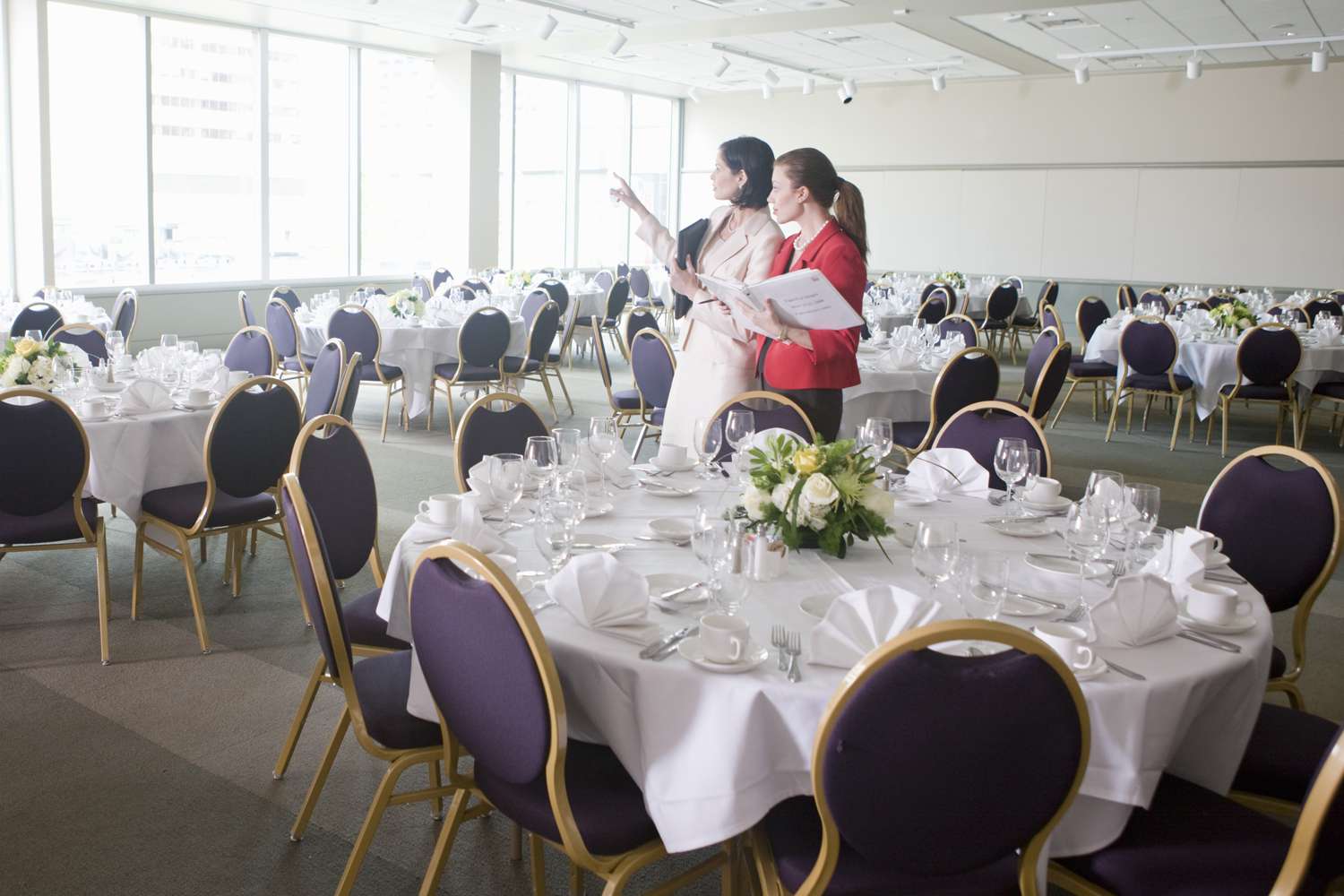 Two event planners discuss ideas while looking at an empty banquet hall with set tables and chairs.