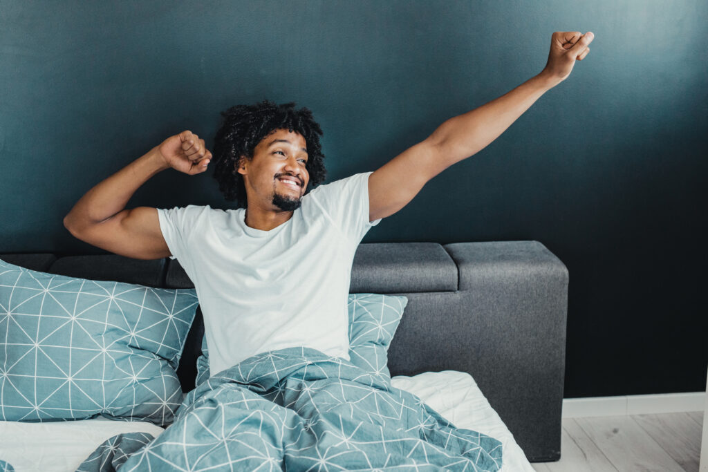 A young man stretches his arms and smiles while sitting up in bed, ready to start his day.