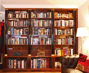 Dark wood bookshelves filled with various books, including hardcovers, paperbacks, and leather-bound books, as well as some knick-knacks and a lamp.