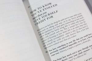 A book opened to a page with a black and white image of a person's hand holding a pen and a magnifying glass over a piece of paper with the words 'It's hard to see how far along the path you've come while you're so focused on taking each step' written on it.