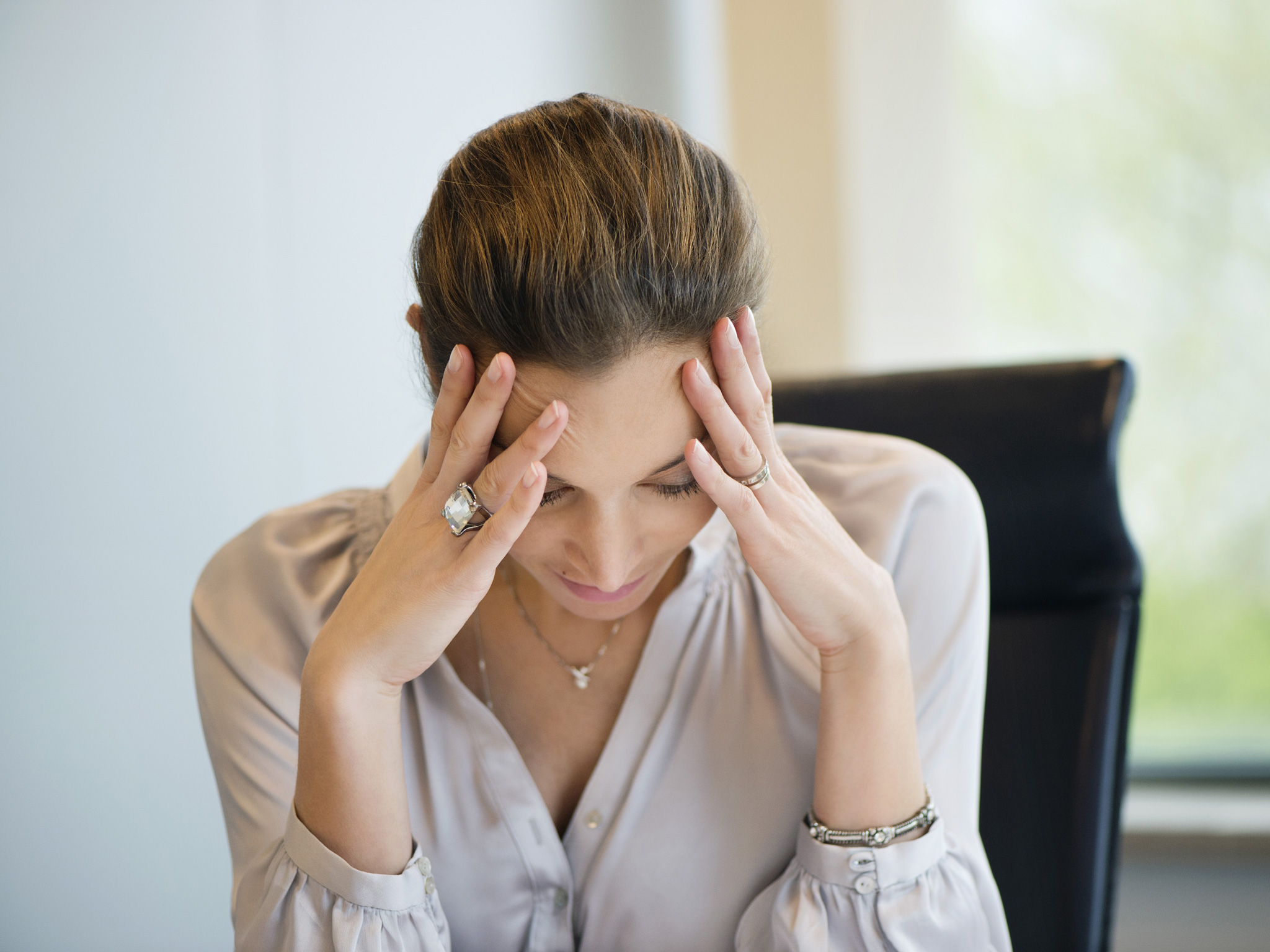 A woman wearing a silver blouse holds her head in her hands while sitting at a desk.