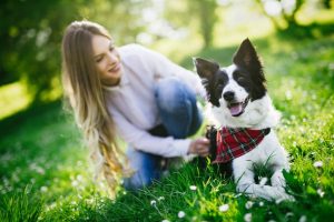 A smiling woman kneels in the grass next to her happy dog.