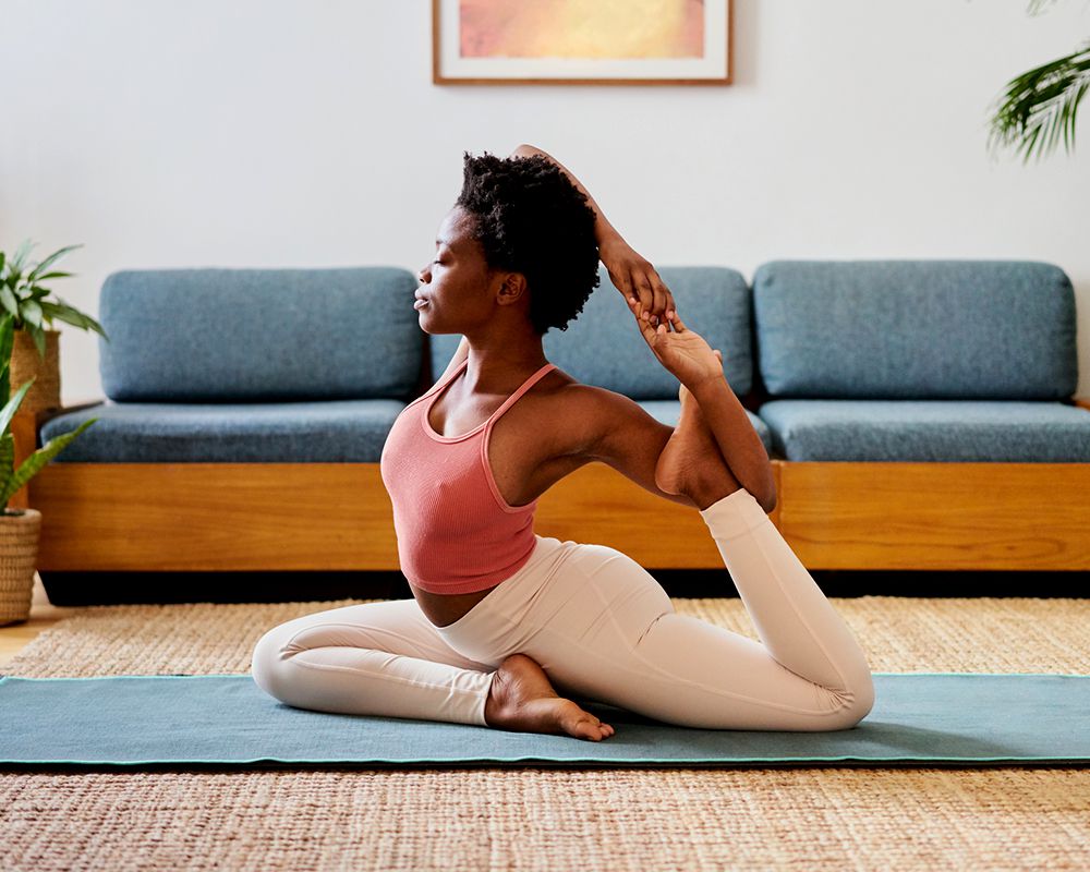 A young woman wearing a pink top and white leggings is doing a yoga pose on a blue yoga mat in her living room.