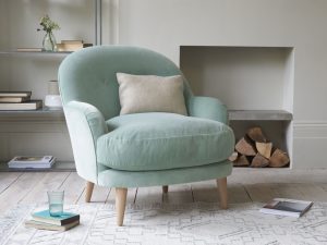 A mint green velvet armchair sits in a cozy study with a gray patterned rug, a stack of books, and a glass of water on the floor next to it.