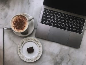 On a marble table is an open silver macbook and a cup of coffee with a small plate with a brownie square on it.