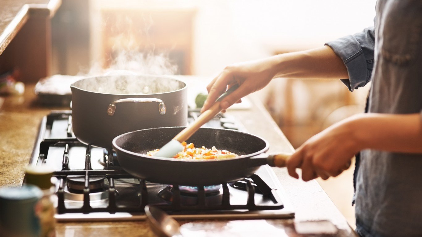 A person is cooking in a kitchen. They are stirring a pan of vegetables with a wooden spoon while a pot of water is boiling on the stove next to it.