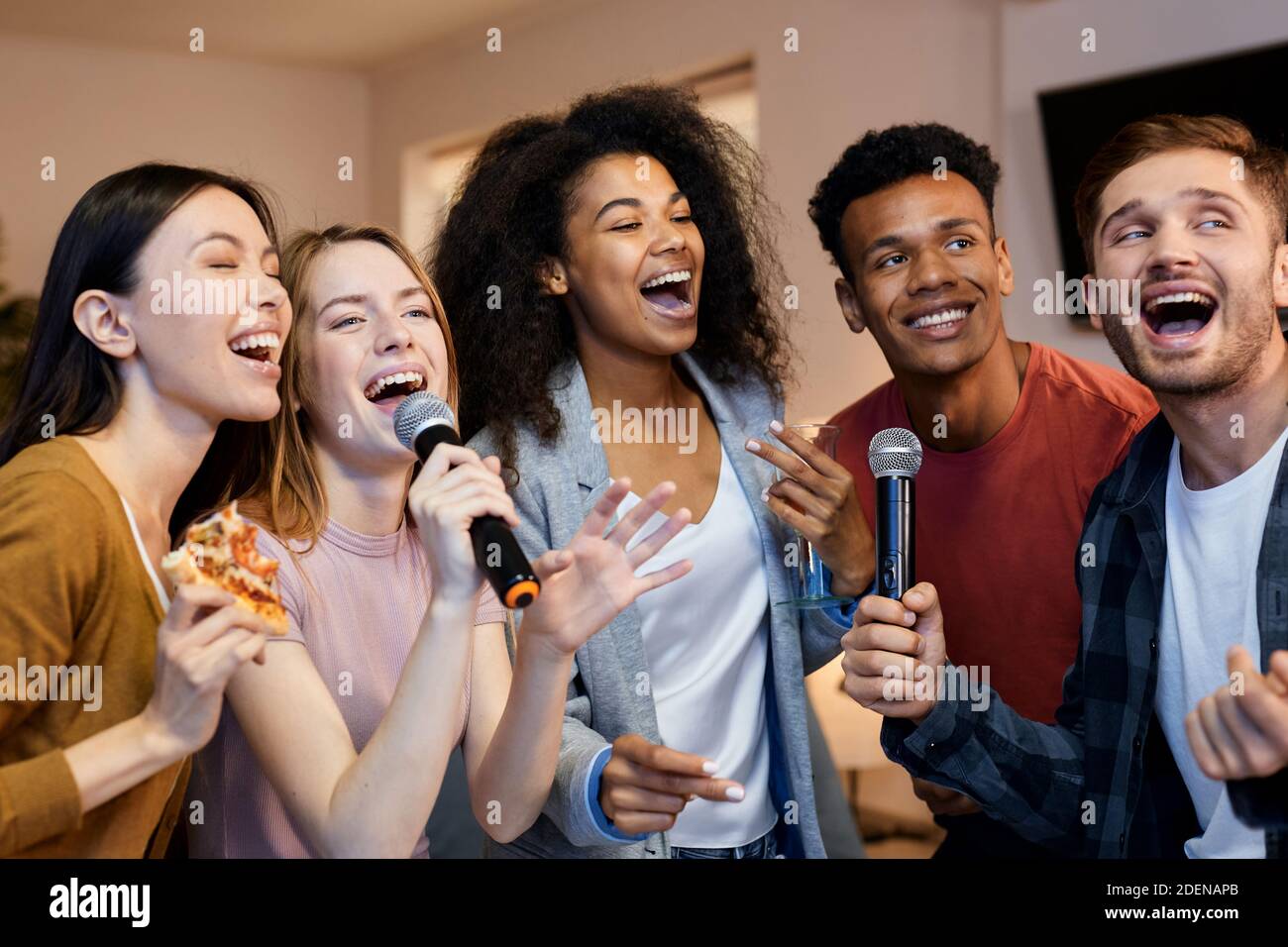 A group of five friends are singing karaoke together, they are all holding microphones and singing with joy.