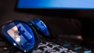 A pair of blue sunglasses with the Facebook logo on the lenses rest on a black keyboard with a blue light in the background.