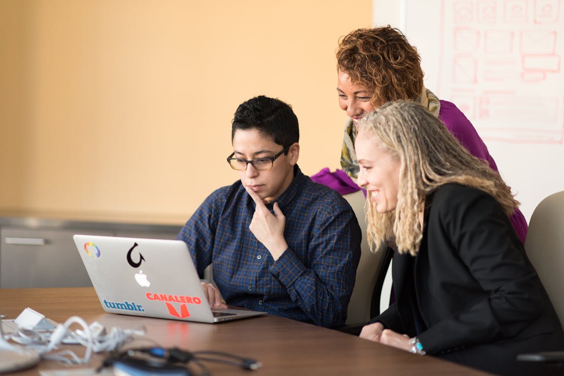 Three people are looking at a laptop. The person in the middle is wearing a blue shirt and has short dark hair. The person to the left is wearing a white shirt with a black and blue flannel shirt over it and has short dark hair with blonde highlights. The person to the right is wearing a black suit jacket with a purple shirt and has long blonde hair.